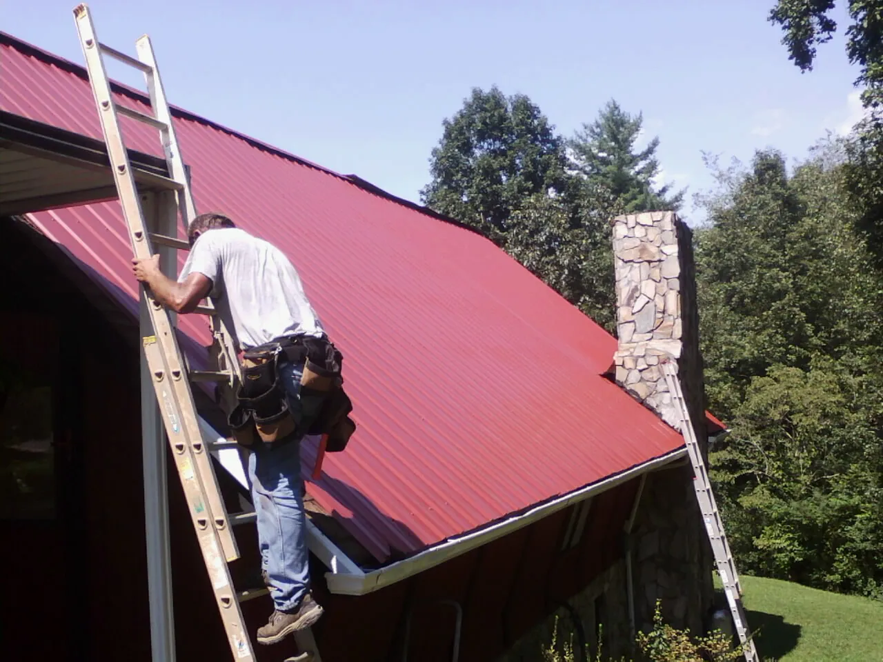 Roofer on a ladder beside a red metal roof and stone chimney — Soffit & Fascia Repair by Roofing Brothers in Welda